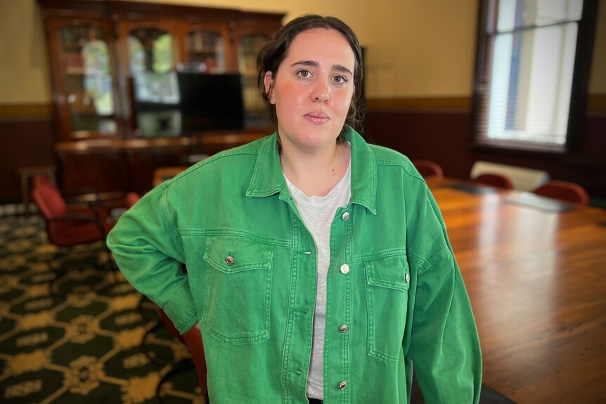 Clare Elliott, in a green bomber jacket & white T-shirt, stands with her right arm resting on her right hip in a heritage room.