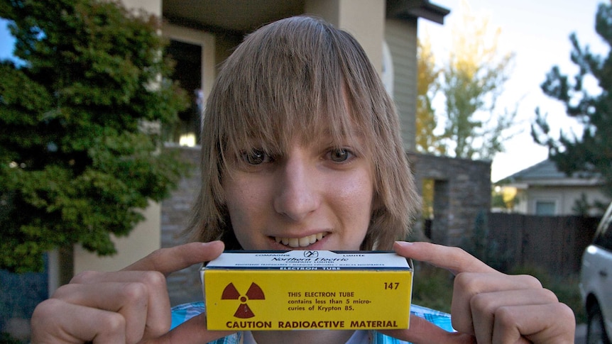 Taylor Wilson looks at the camera while holding up an electron tube box with a radioactive warning label on it