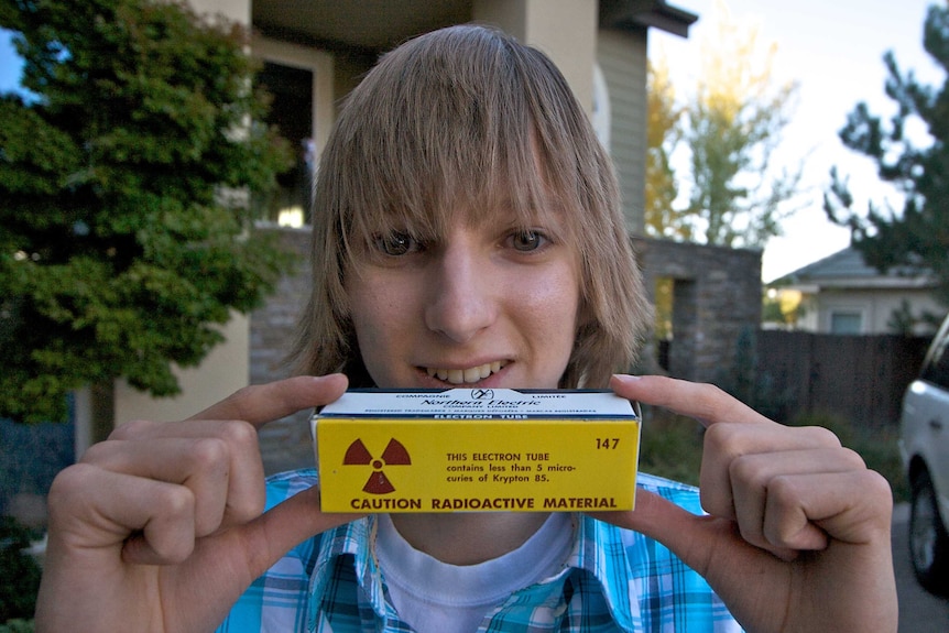 Taylor Wilson looks at the camera while holding up an electron tube box with a radioactive warning label on it