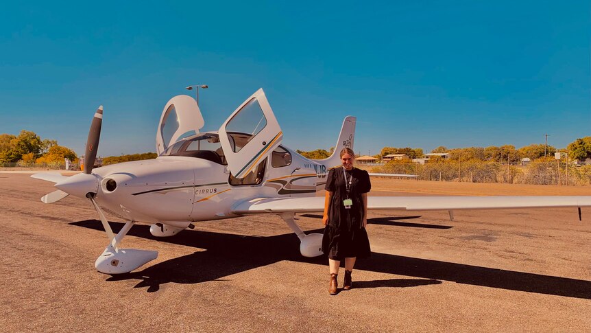 a woman in a black dress stands in front of a small flying doctors plane 
