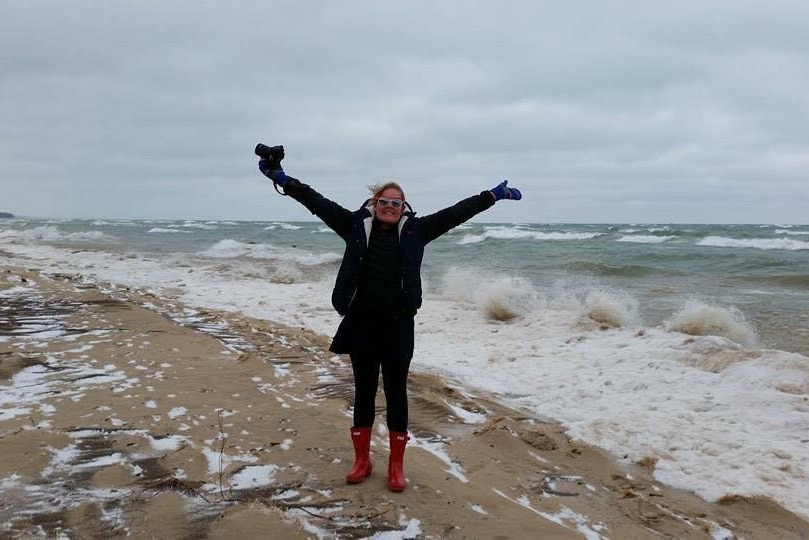Kristyn Keall at the edge of a lake, with snow on the ground, wearing a big jacket and mittens and looking happy.