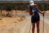 Mina Guli walks up a dirt road in harsh sunlight with headphones in, a cap on and support sticks to aid fractured femur.