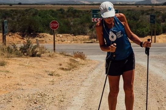 Mina Guli walks up a dirt road in harsh sunlight with headphones in, a cap on and support sticks to aid fractured femur.