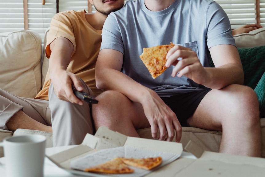two men cuddling and eating pizza on the couch