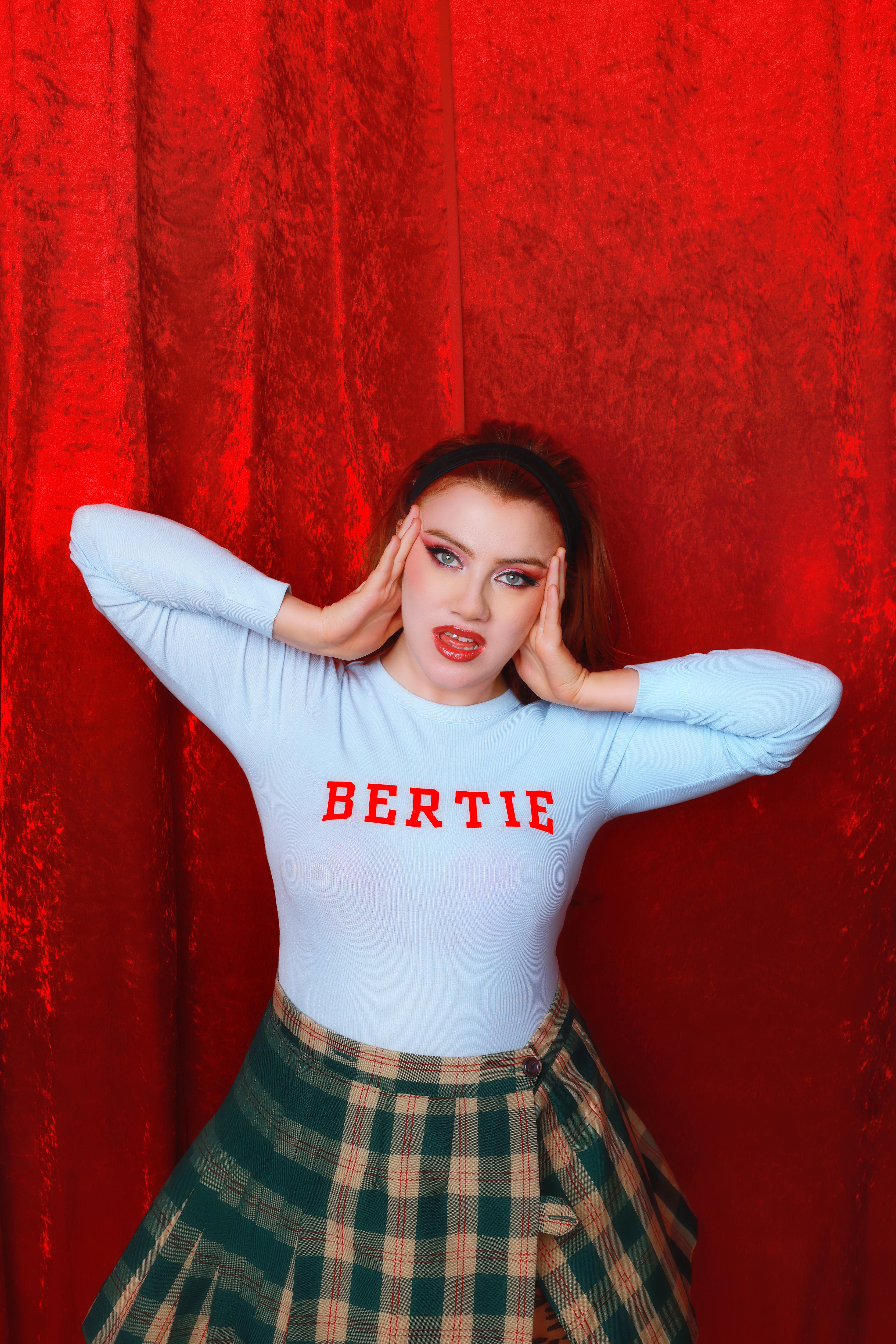 Ciara wears a blue top with the word 'Bertie' on it, a tartan skirt against a red background holding her hands up to her face