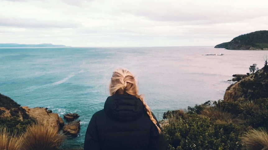 A woman stands looking out at the ocean with a puffer jacket on.