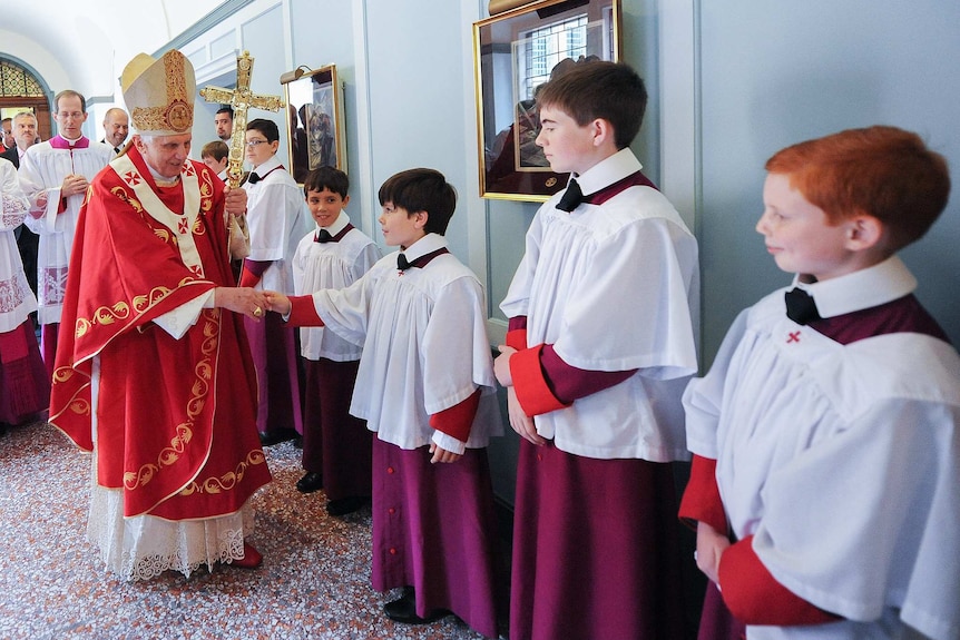 You view Pope Benedict XVI in red ceremonial robes shaking hands of young boys in white and purple vestments.