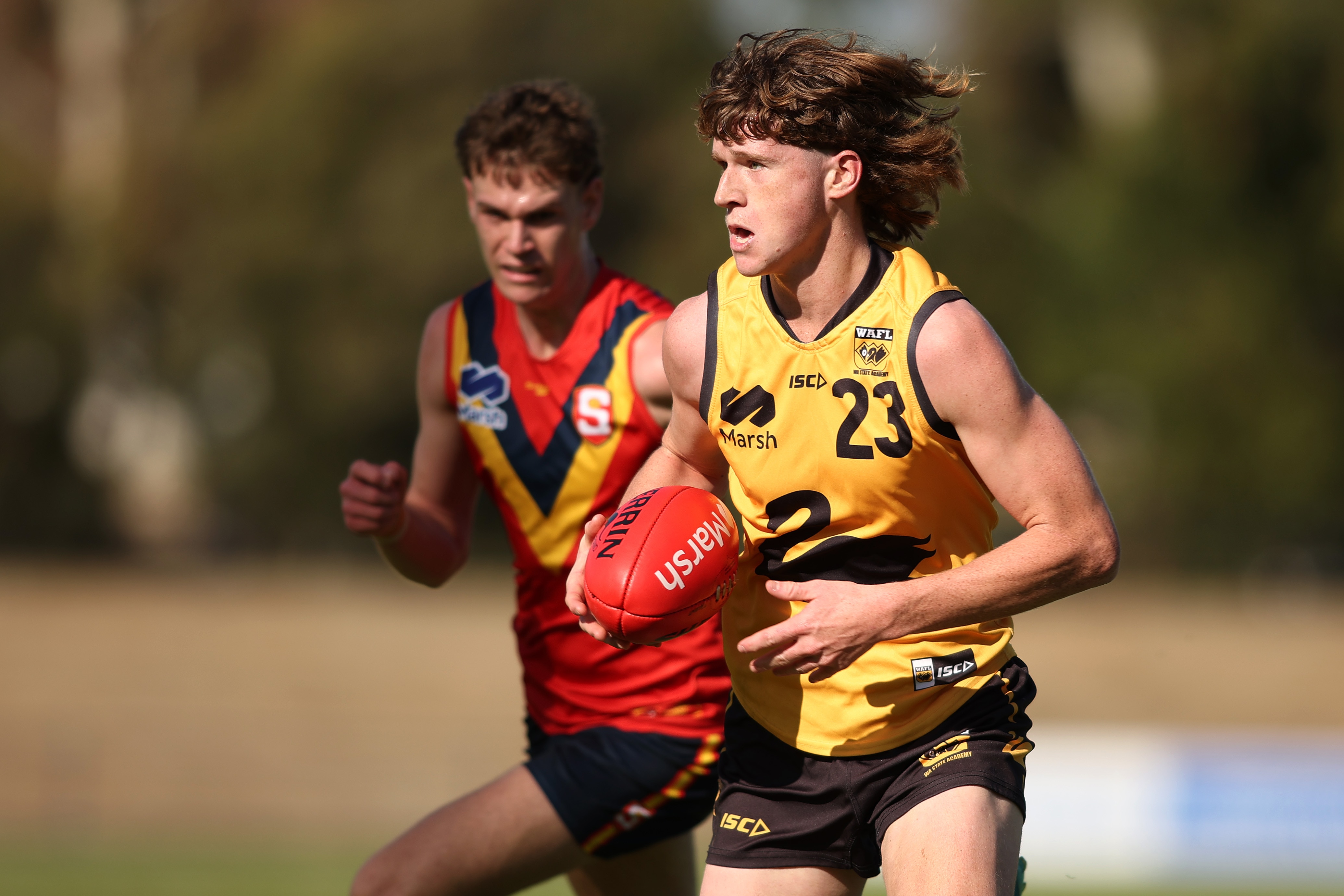 A young footballer with long hair runs while holding the ball and being chased by another footballer.