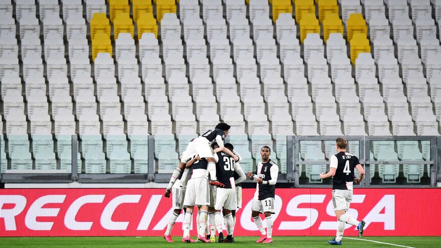 A huddle of players in black and white shirts celebrate in a soccer match in front of empty seats.