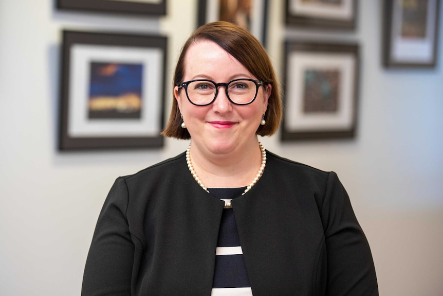 A woman in corporate attire smiles at the camera with picture frames in the background