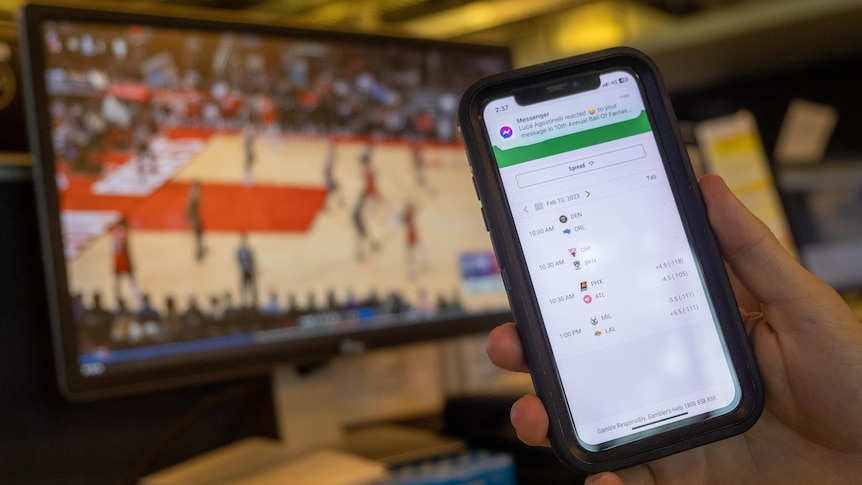 A man's hand holding a phone with betting odds. The phone is receiving a message from a group chat. 