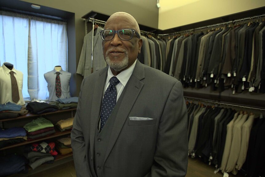 An older African American man standing in a room full of business suits