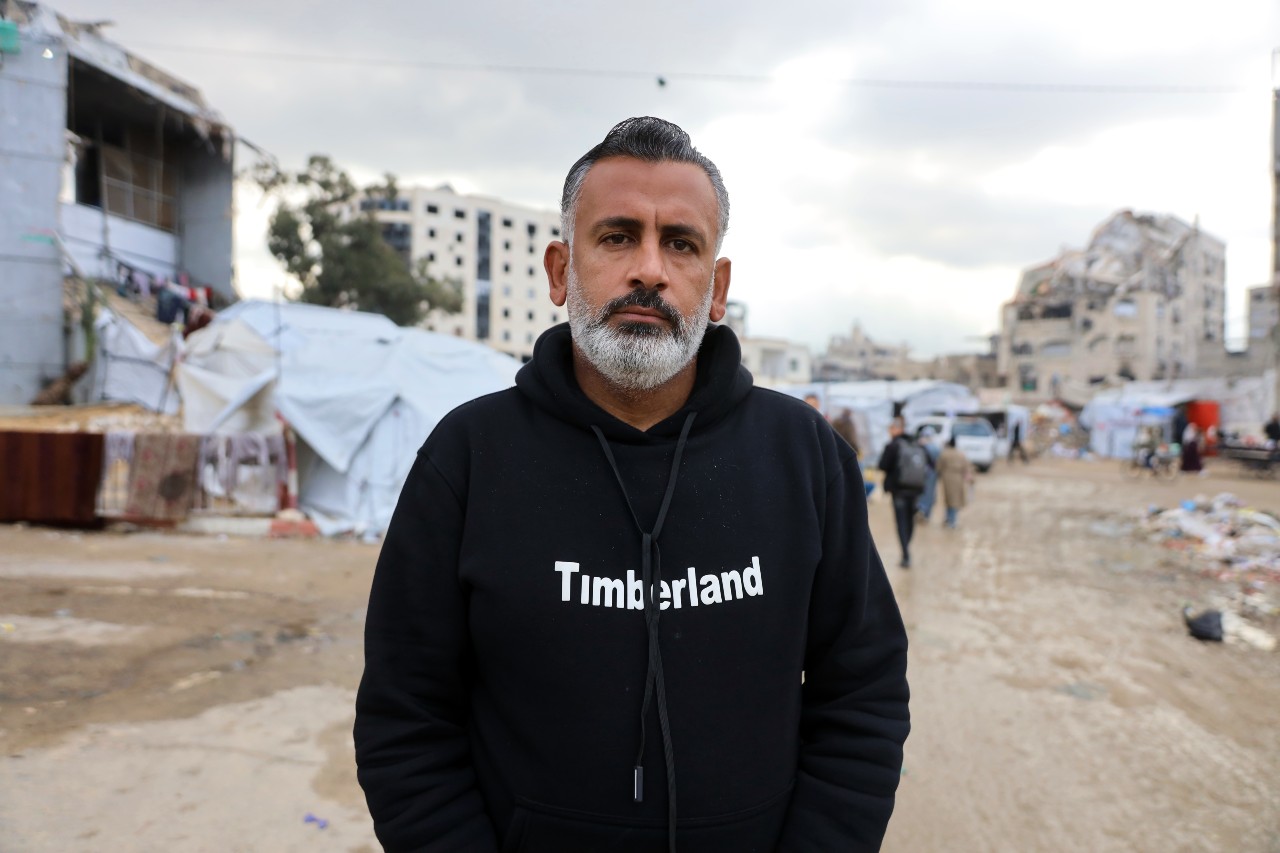 Um homem olha para a câmera com uma expressão neutra. Edifícios danificados visíveis ao fundo.