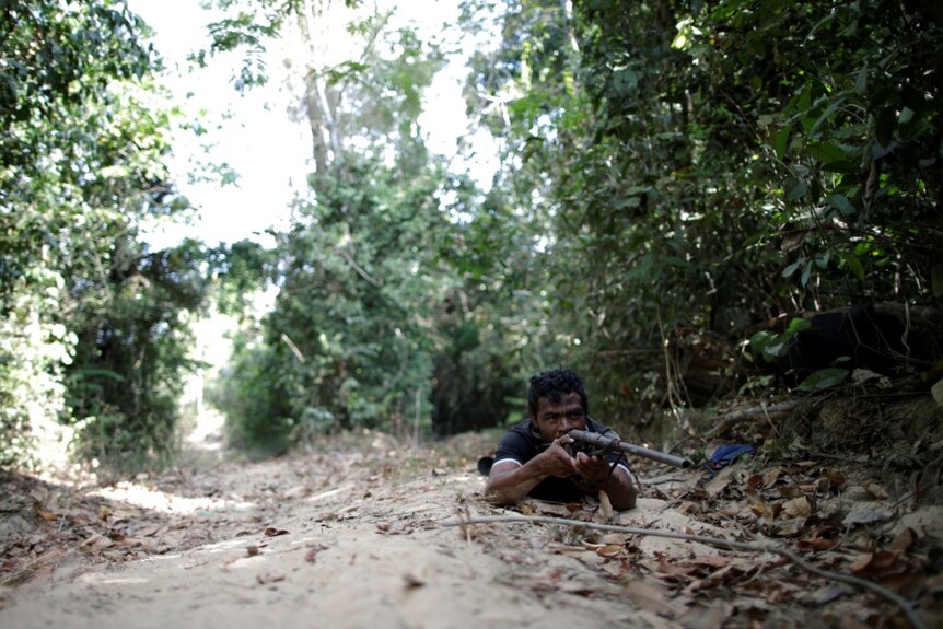 Paulo Paulino lies on his stomach on the Amazon forest floor holding a gun.