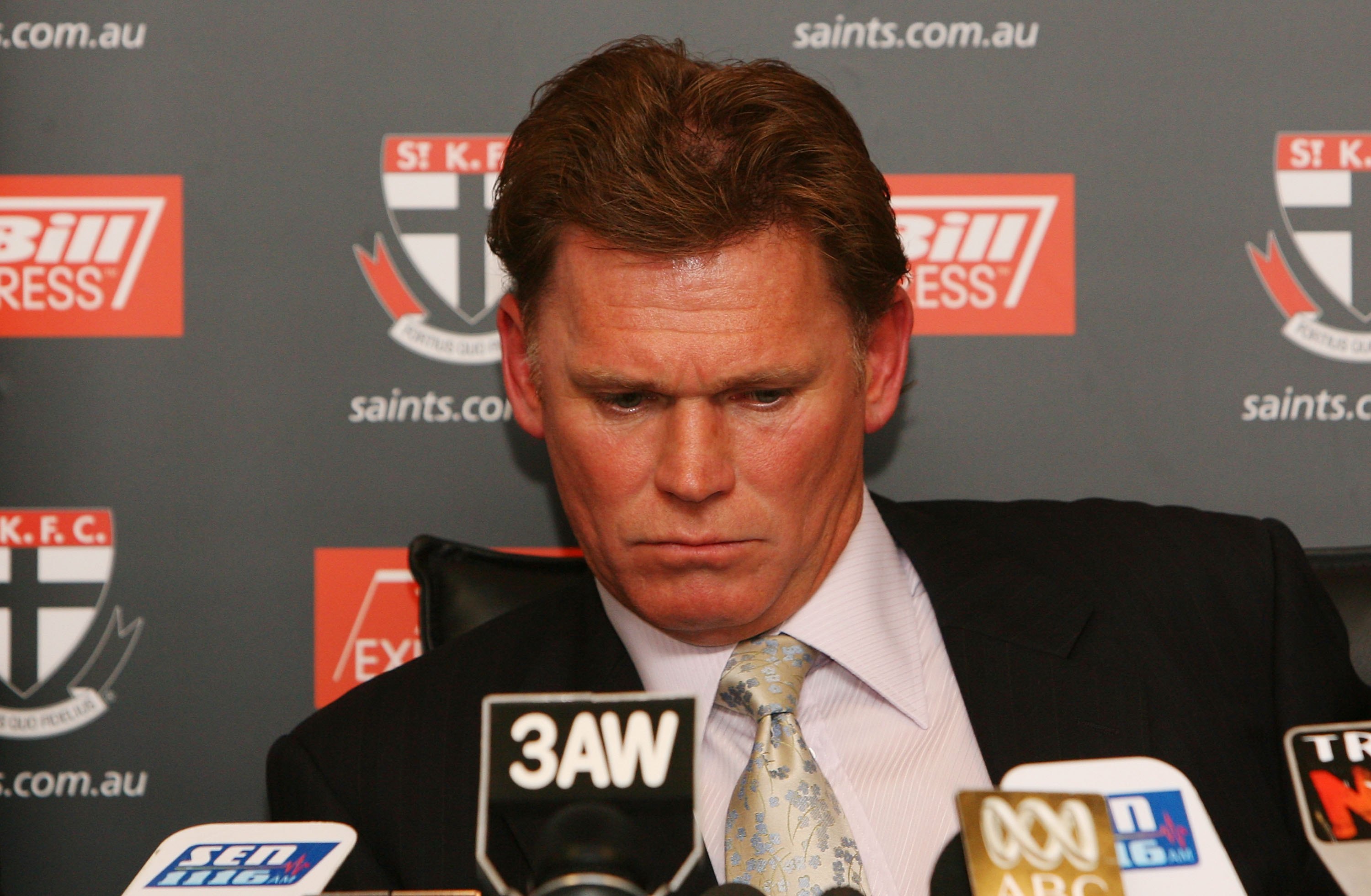 A serious-looking AFL club president looks down as he sits in front of a row of microphones.