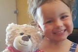 A close-up of a smiling young girl with brown eyes, holding a teddy bear.