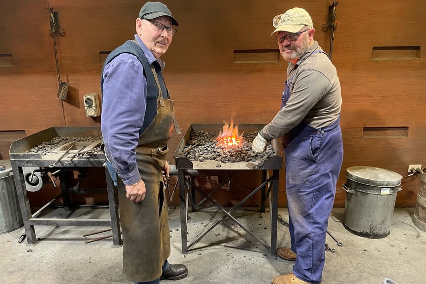 Two men standing in front of a forge looking at the camera with tools in their hand 