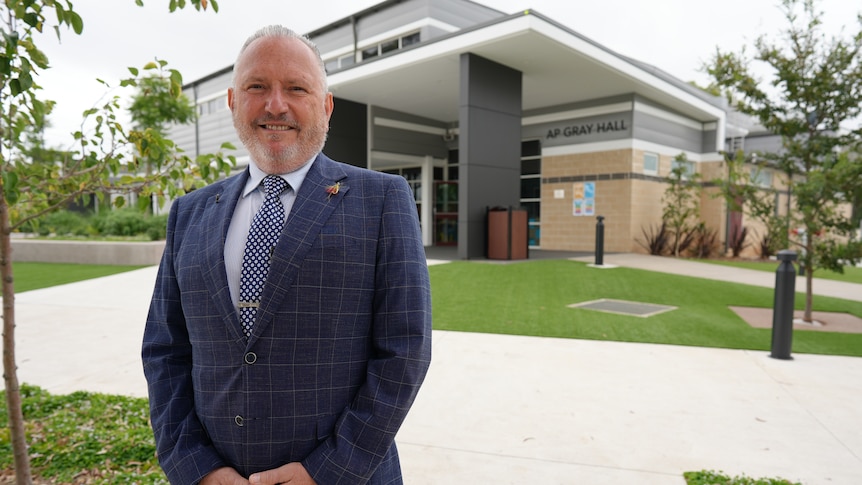 Gabi wearing a blue checkered suit, standing in front of a building at the school.