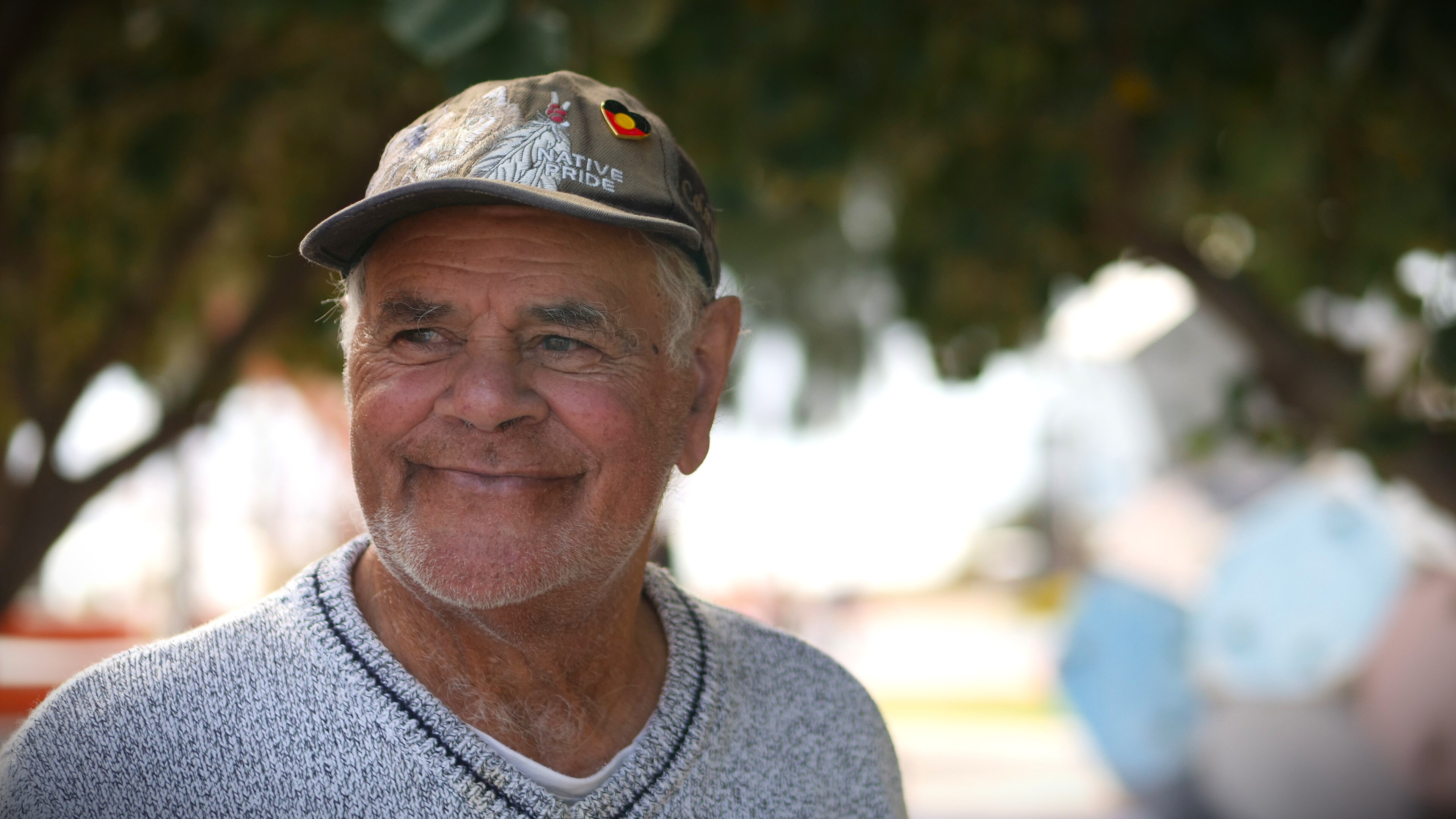 A man in a grey jumper wearing a dark hat smiles past the camera.
