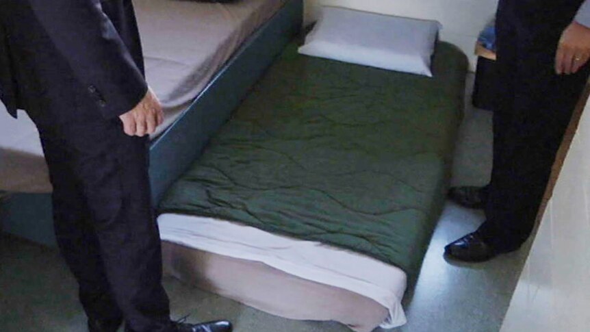 The legs of two men either side of a mattress "double up" on the floor of a prison cell