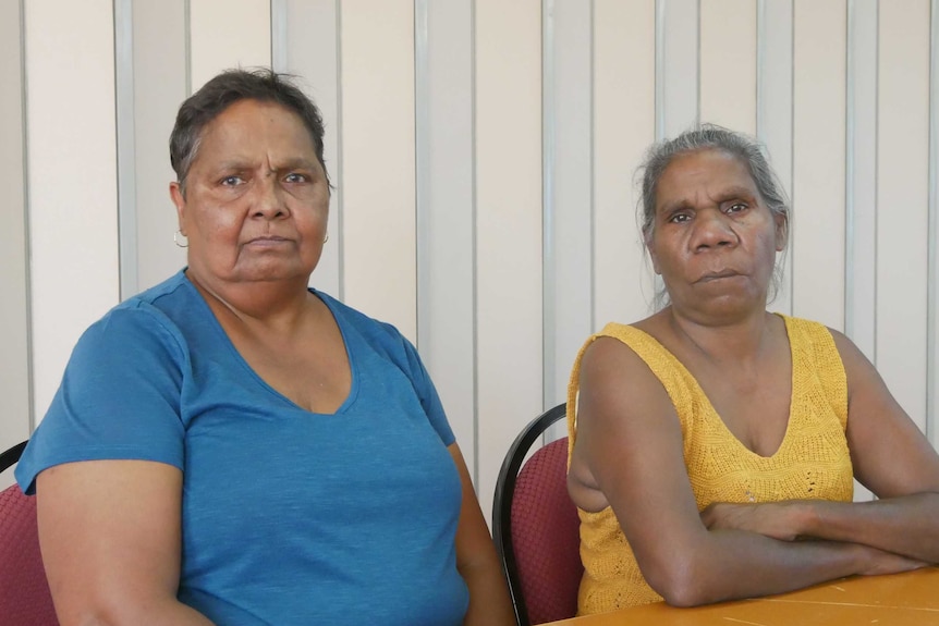 Two women sit at a table. One is wearing a blue t shirt and the other is wearing a yellow singlet.