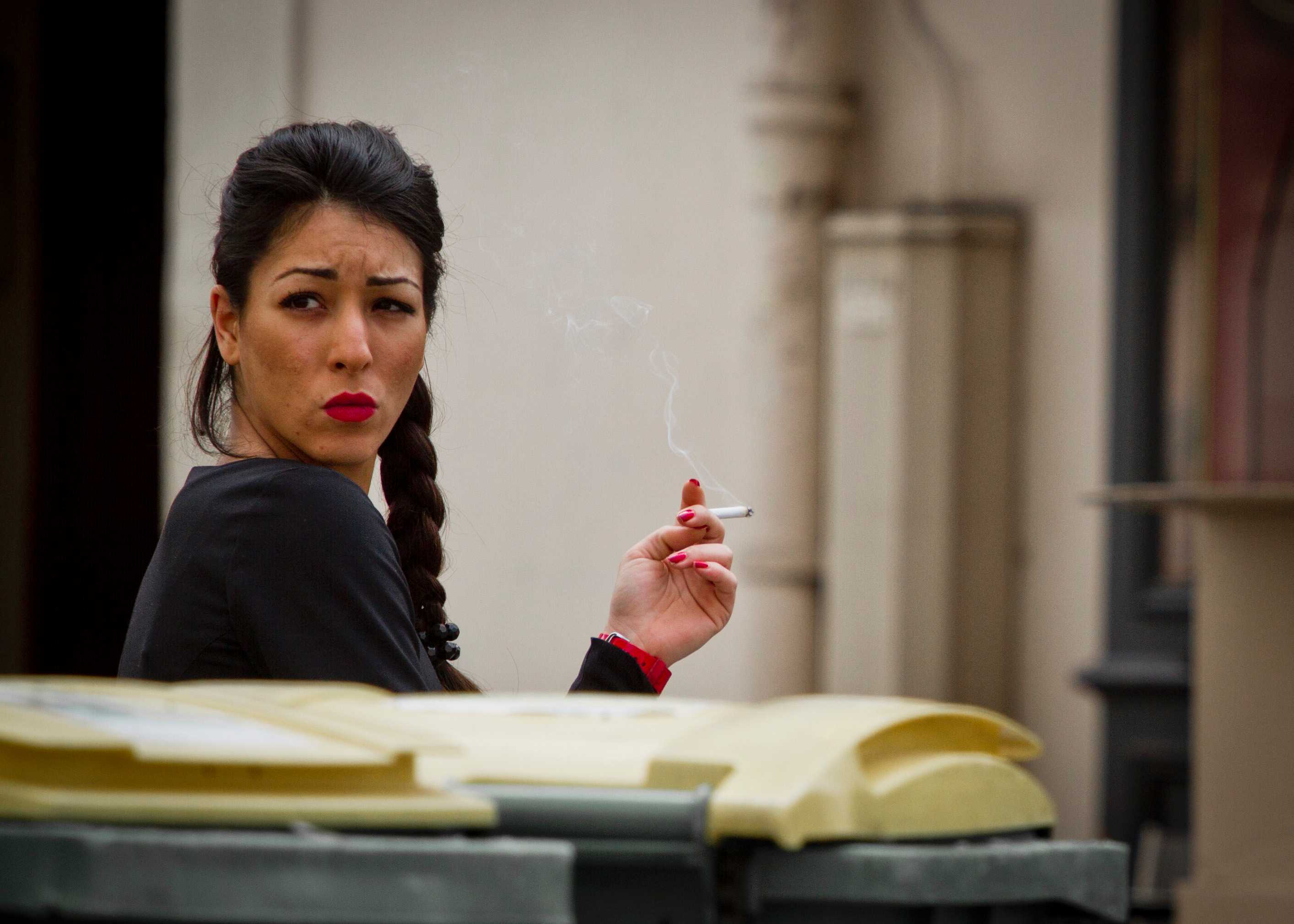A woman standing behind some bins, smoking a cigarette