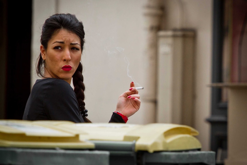 A woman standing behind some bins, smoking a cigarette