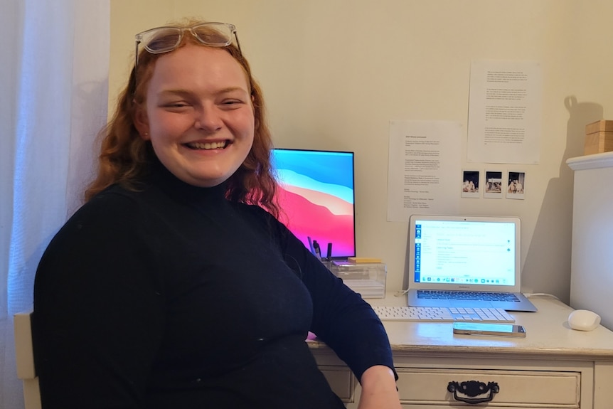 An image of Grace smiling, sitting in front of a white desk, with her back to two screens and wall with documents pinned to it