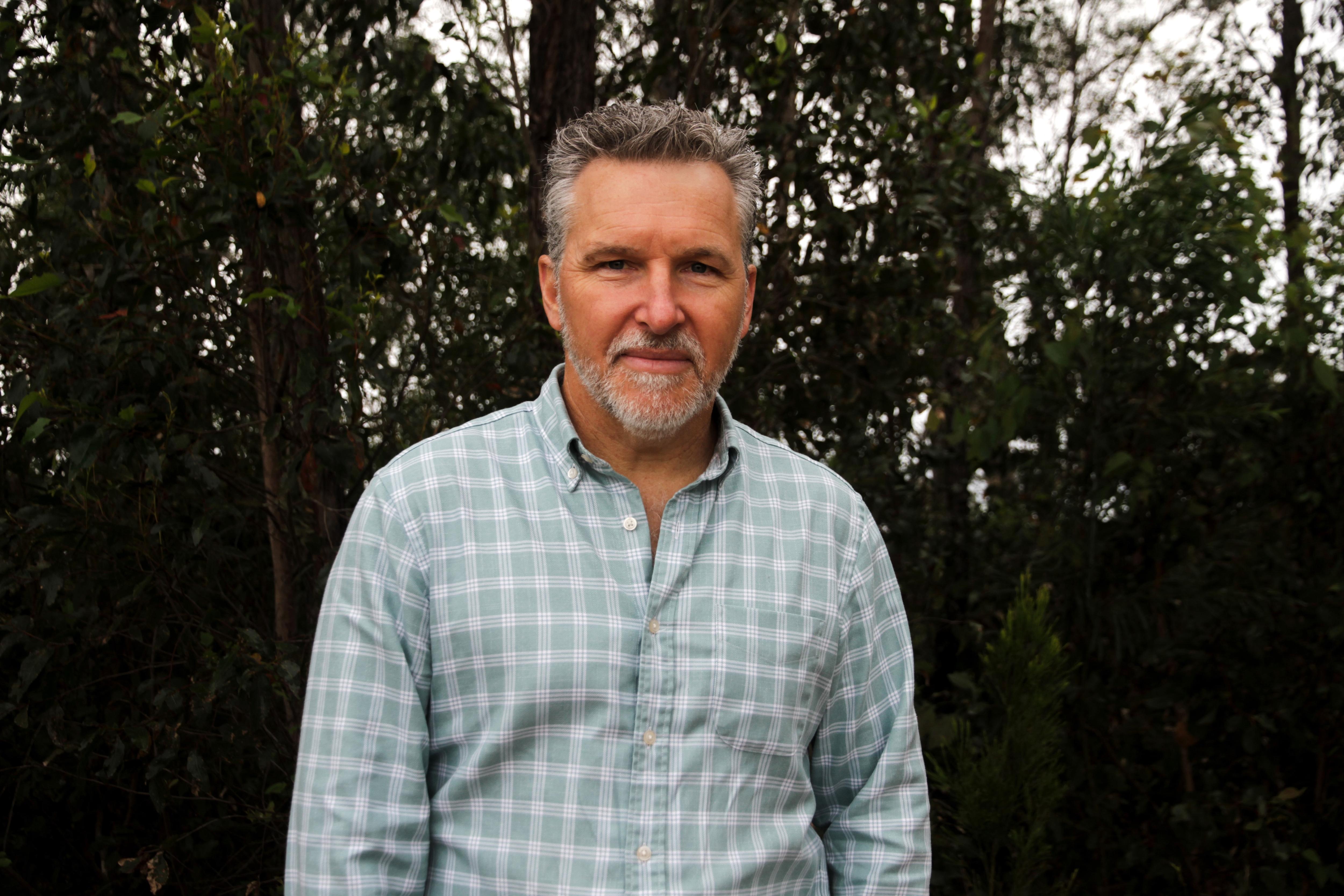 Man in checkered shirt stands in front of scrub