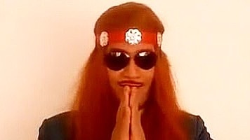 A man with long red hair and wearing sunglasses holds his hands palms facing in front of his face.