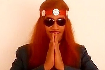 A man with long red hair and wearing sunglasses holds his hands palms facing in front of his face.