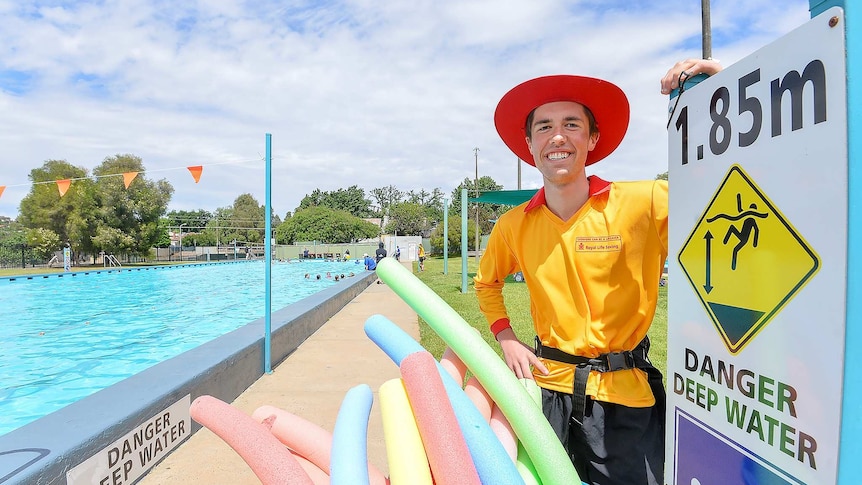 A smiling man of indeterminate age, wearing lifesaver gear and standing in front of a pool.