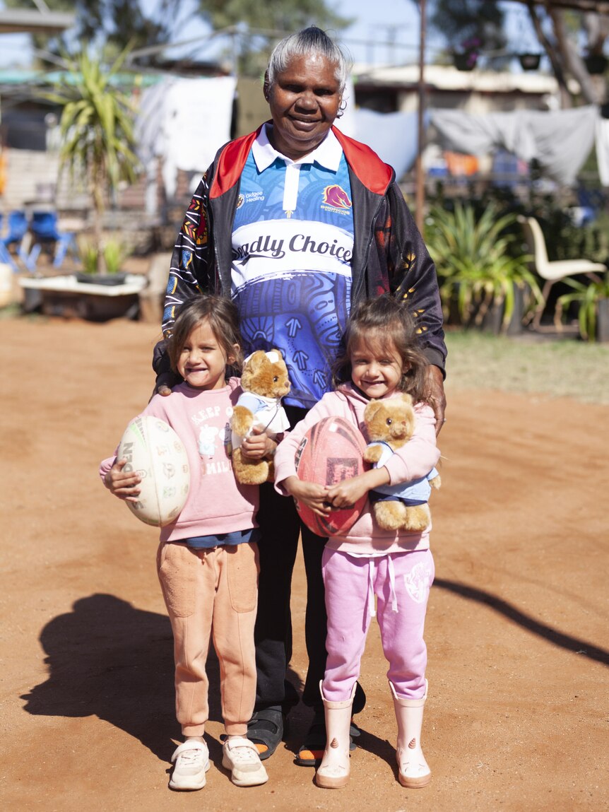 An older Aboriginal woman stands outside with two small toddlers on either side of her smiling.
