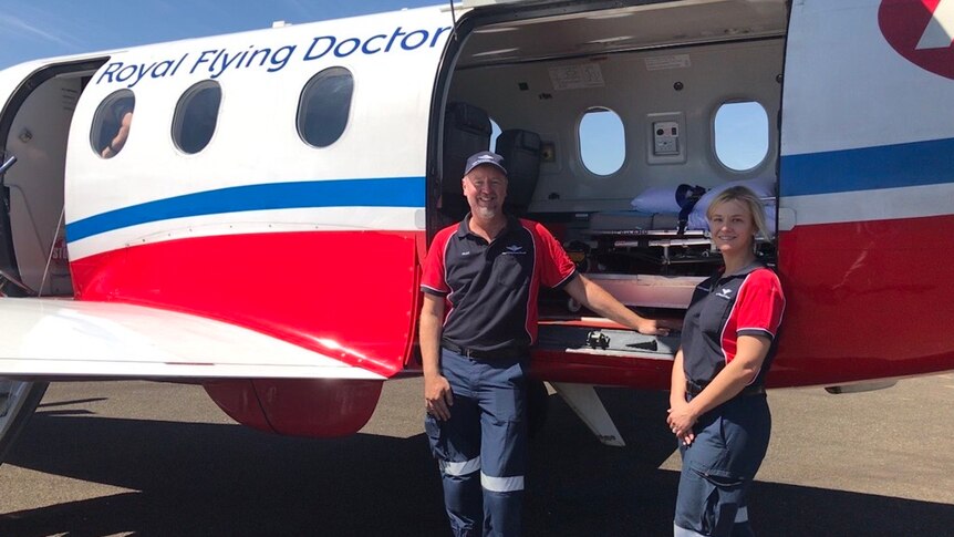 A man and a woman stand outside a Royal Flying Doctor Service plane, wearing blue and red shirts.