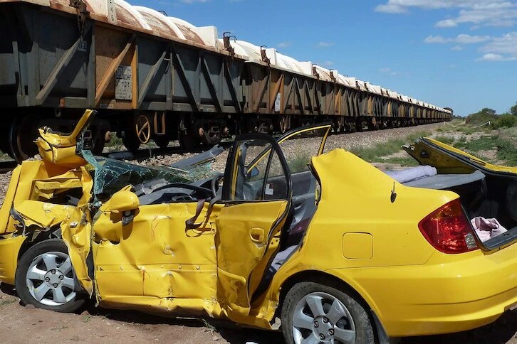 A badly damaged yellow car beside a rail line with train carriages on it.
