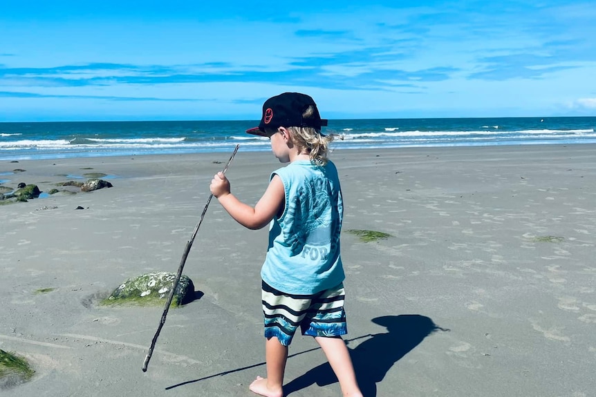 A little boy is walking on the beach with a stick.