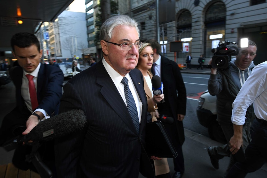 A man with grey hair wearing glasses walks along a street with journalists and camera operators around him.