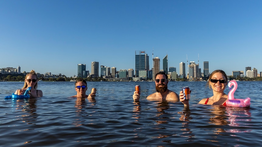 Four people seen from shoulders up in a large body of water, smiling and holding drinks, with skyscrapers on horizon behind.