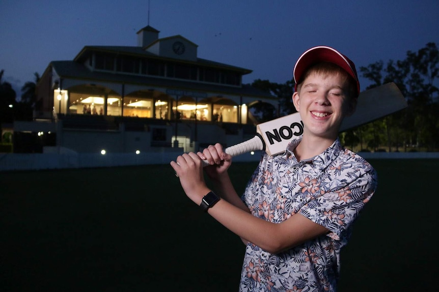 Sean Kendrick holds a cricket bat over his shoulder while standing  on an oval at night with a lit clubhouse seen behind him
