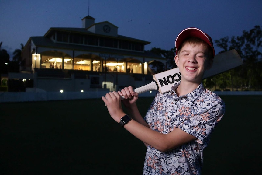 A boy holds a cricket bat over his shoulder stands on an oval at night with a lit clubhouse seen behind him