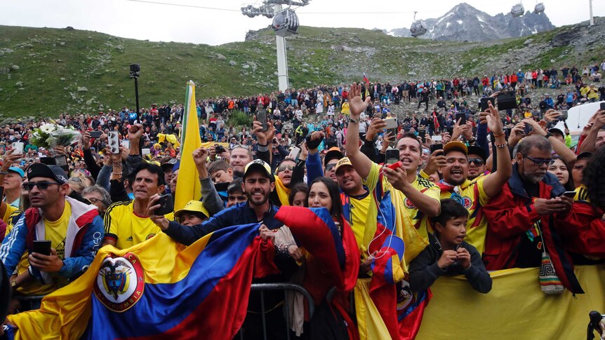 A crowd of people holding Columbian flags cheer on the side of a mountain