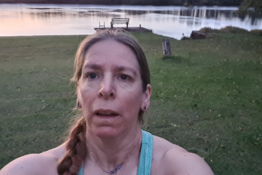 A woman takes a selfie in front of flood water with a seat in the background.