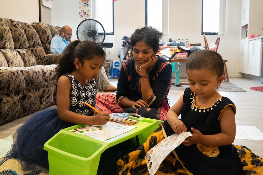 A woman sits between two young girls as they work on a drawing. 