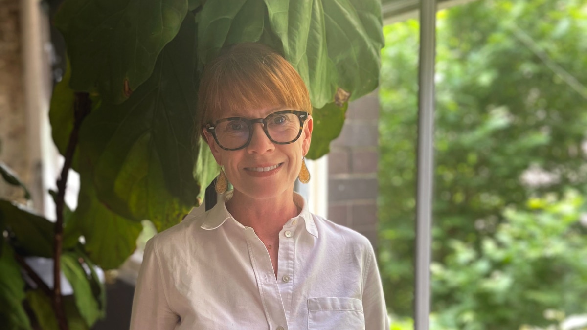 Una mujer de mediana edad con cabello rubio fresa y gafas negras con una camisa blanca sonríe a la cámara en una habitación