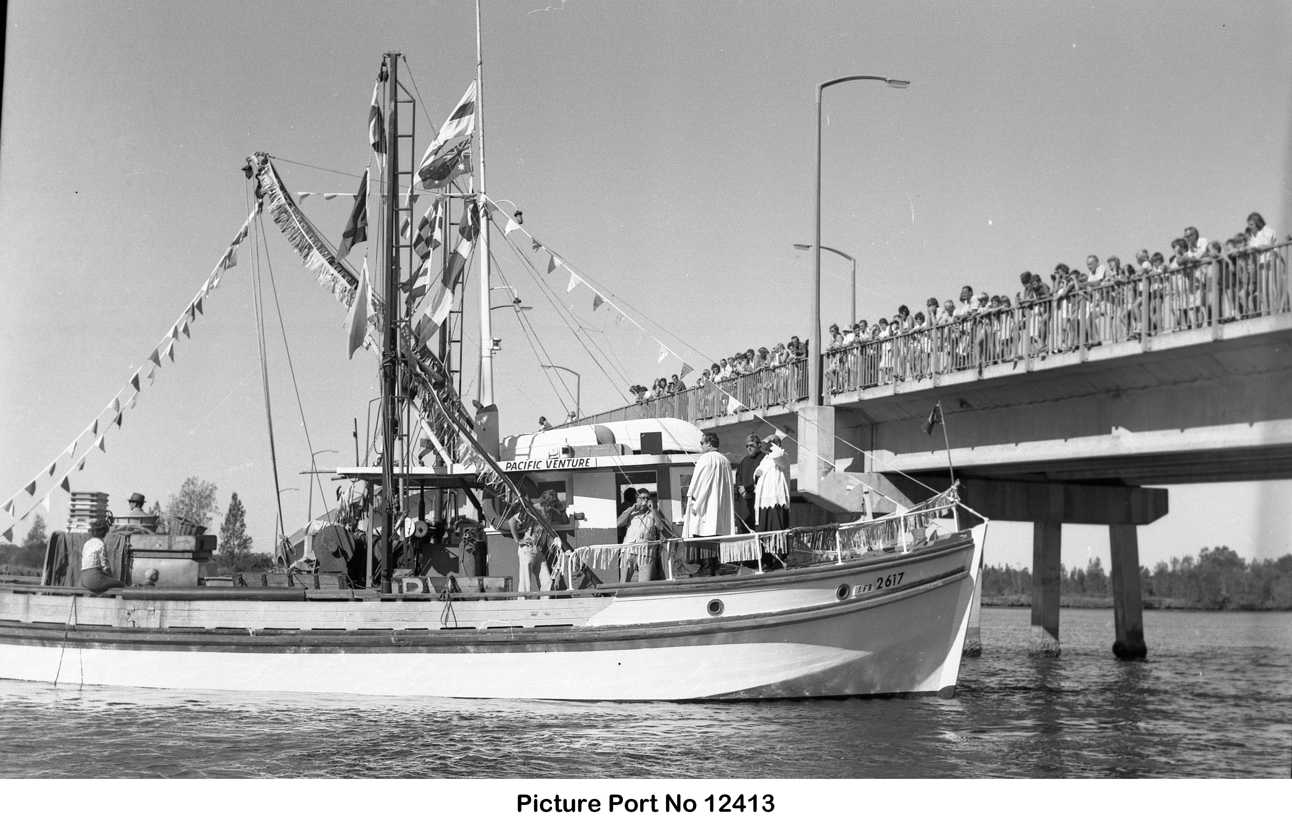 A black and white image of an old fishing boat near a bridge with people lined along the bridge.