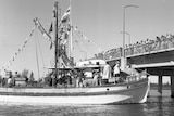 A black and white image of an old fishing boat near a bridge with people lined along the bridge.