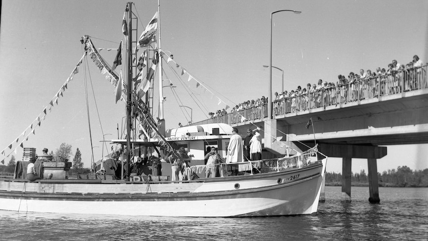 A black and white image of an old fishing boat near a bridge with people lined along the bridge.