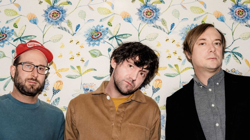 The three members of American band Bright Eyes stand in front of floral wallpaper.