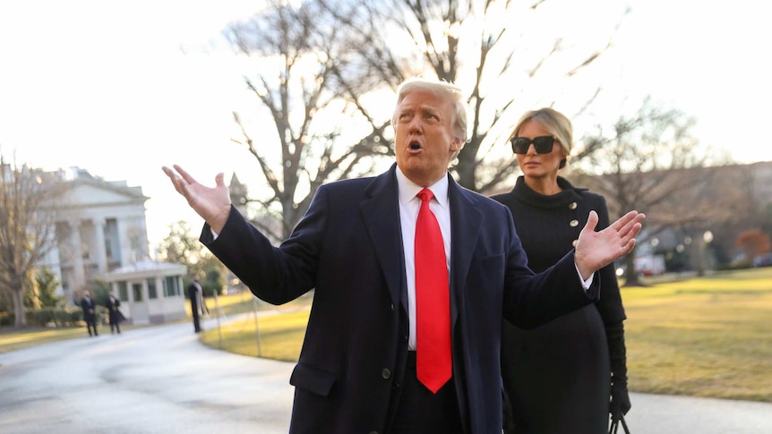 Donald Trump wearing a suit with red tie spreads his hands in the air next to Melania Trump wearing black with sunnies