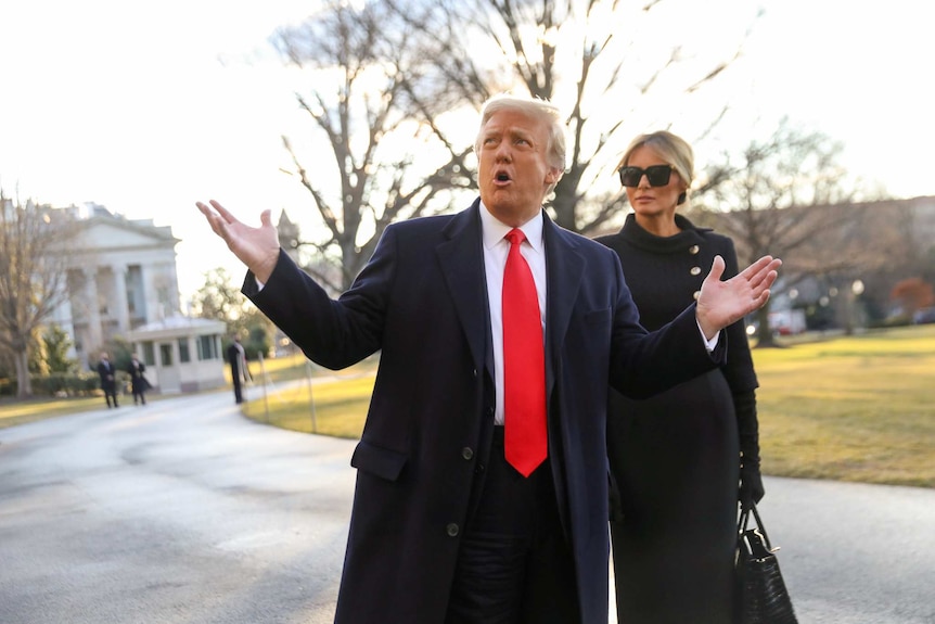 A man with white hair wearing a suit with red tie spreads his hands in the air next to a woman wearing black with sunnies